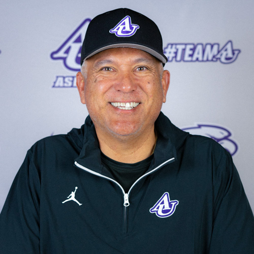 Male Coach profile headshot in a gray polo outside the front of a school blurred with text over his face.